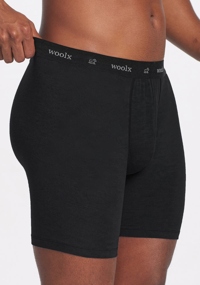A person is shown from mid-thigh to waist wearing Woolx Jazzy No Fly Boxer Briefs in black, made of soft Merino wool and featuring a "woolx" waistband in white text, against a plain background.