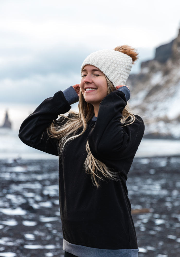 A woman wearing the Kaylee Beanie in cream from Woolx, along with a black sweater, smiles with her eyes closed while standing on a snowy beach. The sky is cloudy, and icy cliffs are visible in the background.