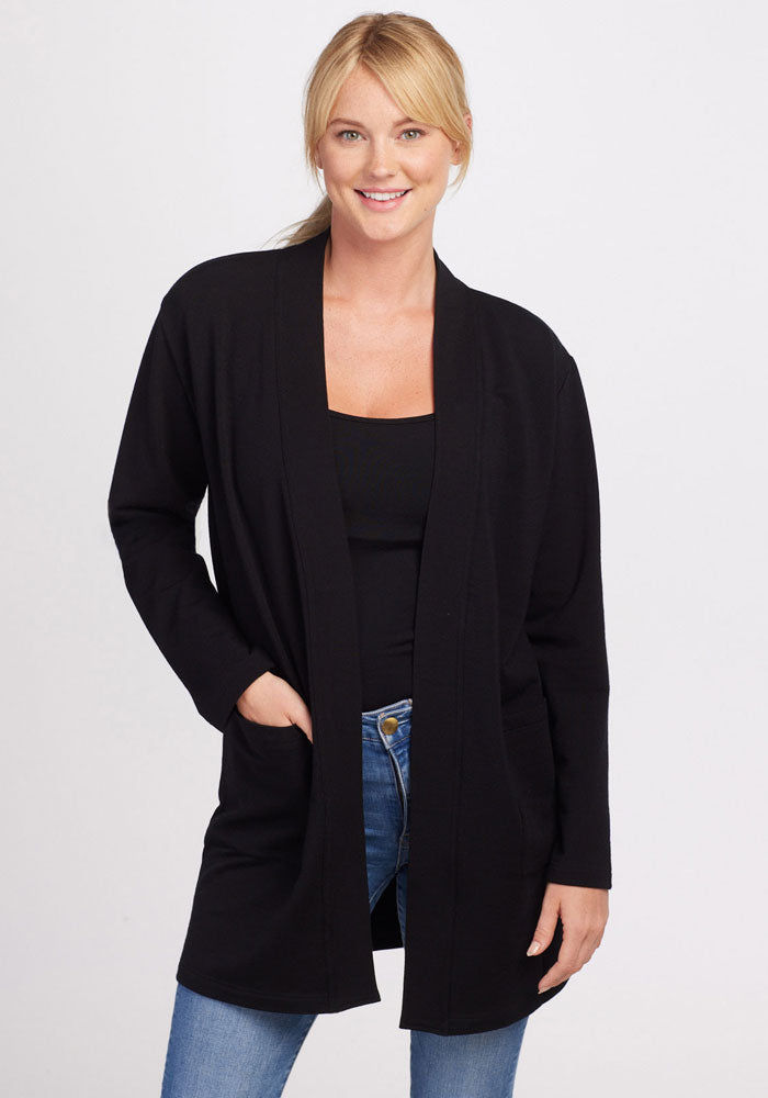 A person with blond hair is smiling at the camera, standing against a plain white background. They are wearing the Woolx Kinsley Cardigan in black over a matching black top and blue jeans. One hand is placed in the pocket of the cardigan, while the other hand rests by their side. | Karly is 5'10", wearing a size S