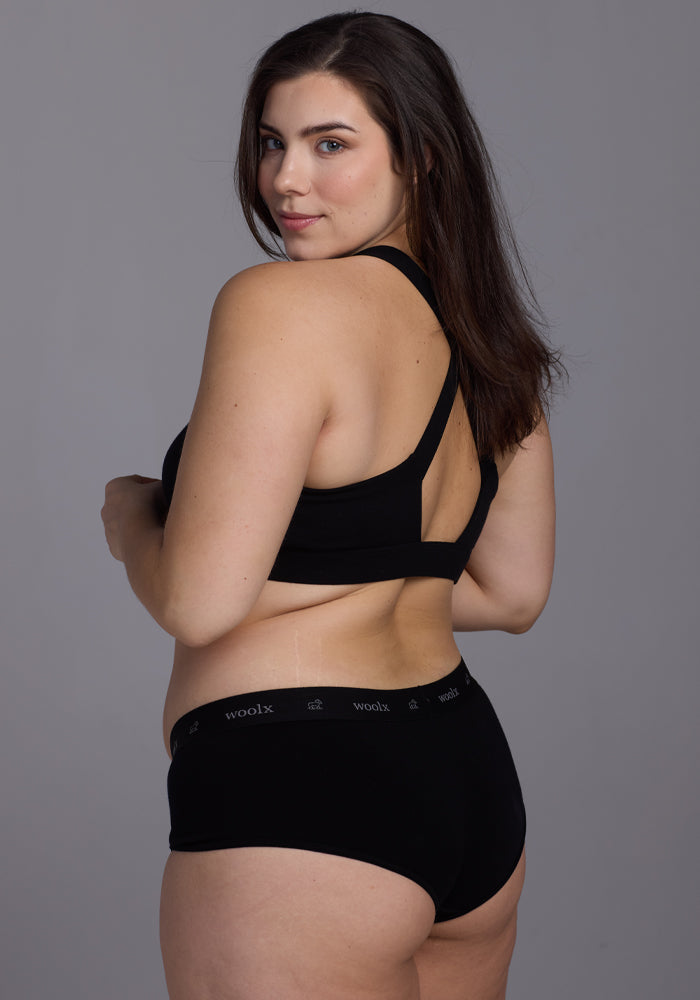 A person with long dark hair, dressed in a Woolx black sports bra and Lila Boy Shorts, stands against a gray background, smiling over their shoulder.