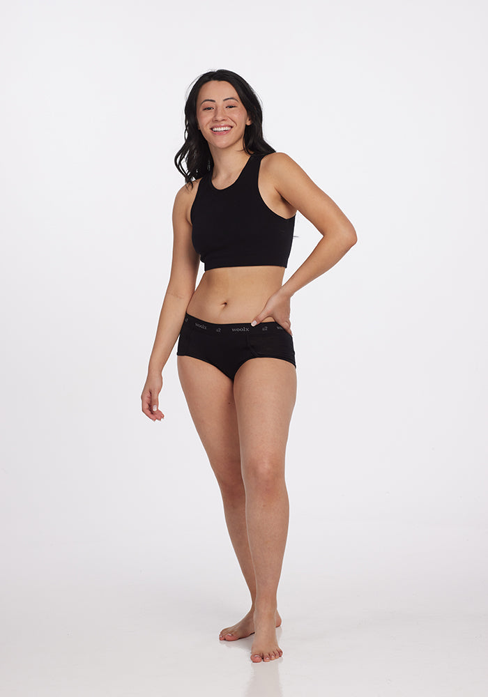 A person stands smiling against a plain white background, wearing a black sleeveless crop top and the Lila Boy Shorts by Woolx, crafted from lightweight fabric. They are barefoot with long, dark, wavy hair.