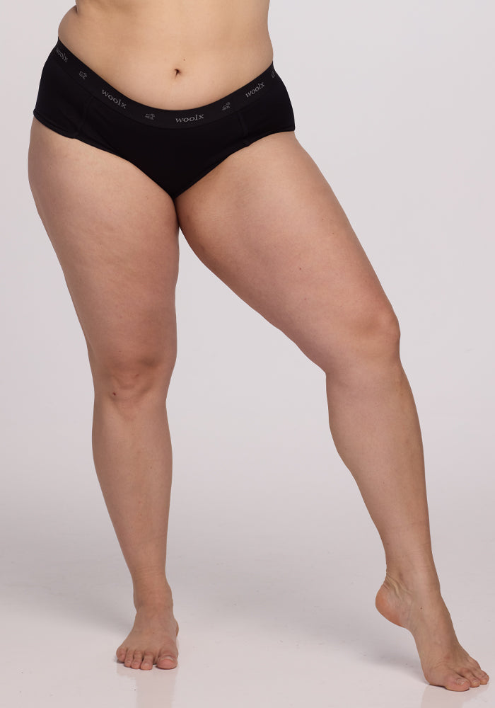 A person is seen wearing Woolx Lila Boy Shorts in black Merino wool, standing barefoot against a simple backdrop. The visible lower half highlights the natural and comfortable fit.