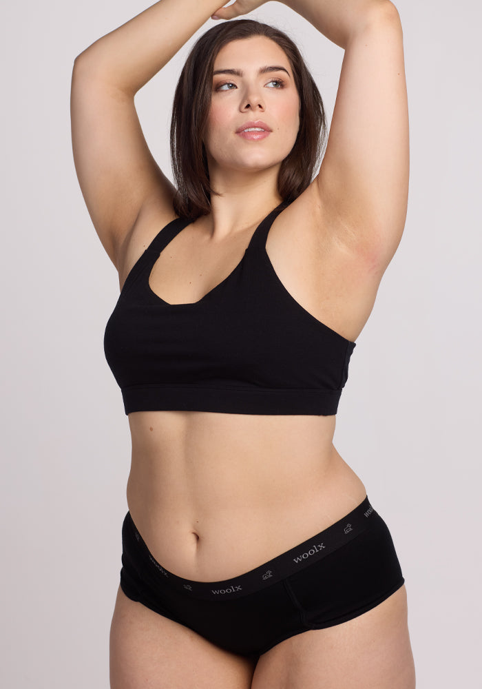 A dark-haired woman poses confidently wearing a black sports bra and Lila Boy Shorts - Black by Woolx, with "woolx" on the waistband. Made from soft Merino wool for a comfortable fit, she raises her arms against a neutral backdrop.