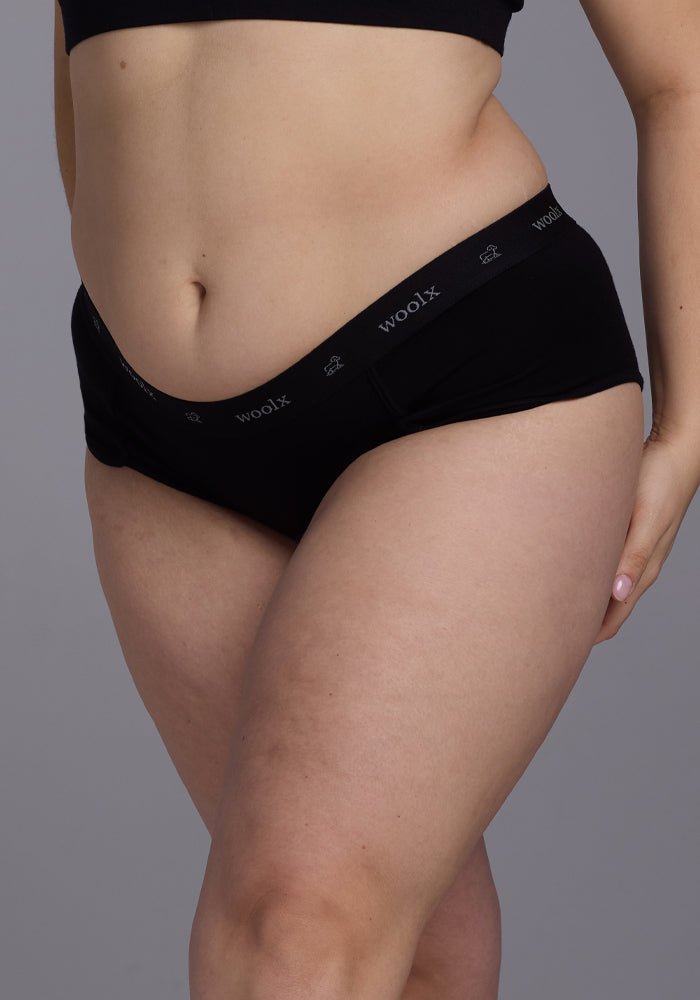 A person wearing black Lila Boy Shorts by Woolx, which are noted for their Comfort Fit, with a waistband showing the brand name in white text against a plain gray background.