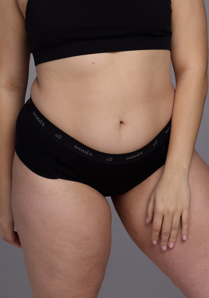 A person wearing the Woolx Lila Boy Shorts - Black confidently poses against a gray backdrop. The focus on the midsection emphasizes the merino wool fabric, ensuring a comfortable fit that combines style and practicality.