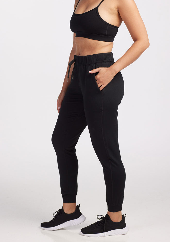 A person wearing a black sports bra, Woolx Lola Jogger in black, and black sneakers is standing with one hand in a pocket against a white background. Their face is not visible in the image.