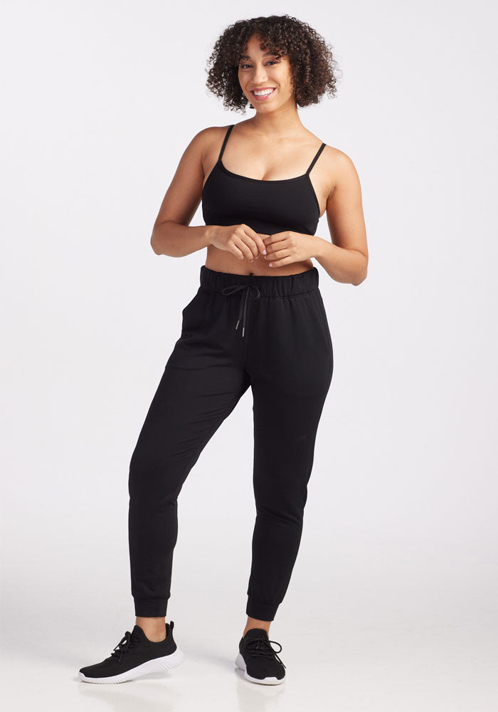 A woman smiles, standing confidently in a black sports bra, Woolx's Lola Jogger - Black in Merino wool, and black sneakers. The odor-resistant joggers promise travel-ready comfort. Hands resting near her waist, she poses naturally against a plain white background.