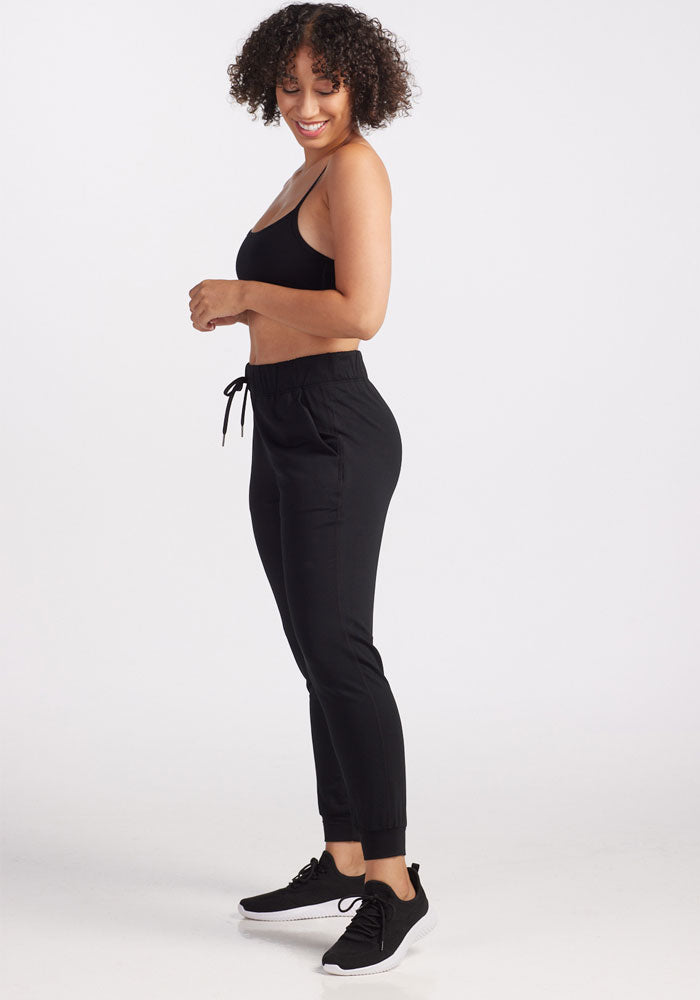 A person with curly hair smiles while standing sideways, exuding travel-ready comfort. They are wearing a black sports bra, stylish Lola Jogger - Black from Woolx Merino wool joggers, and sleek black sneakers. The background is plain white.