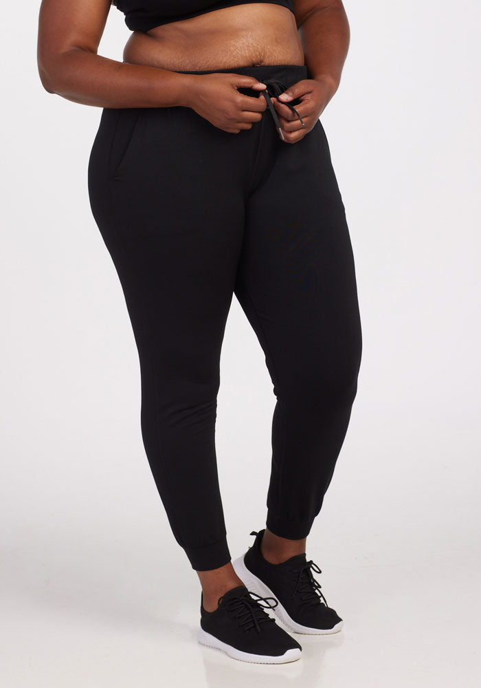 A person dressed in a black crop top and Woolx's Lola Jogger in black, renowned for its odor-resistant Merino wool, is adjusting the waistband. They are also wearing black and white sneakers in a plain white studio setting, perfect for highlighting travel-ready comfort.