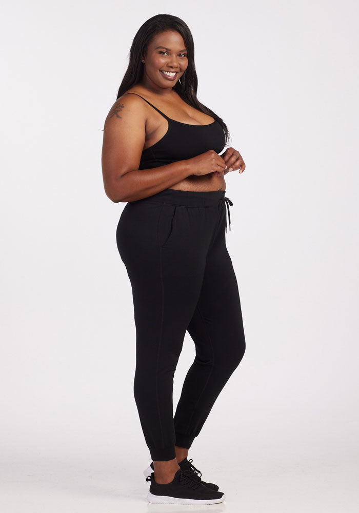 A person is smiling while standing in a side pose against a plain background, exuding travel-ready comfort. They are wearing a black tank top, Woolx's Lola Jogger in black known for its odor-resistant properties, and matching black sneakers.