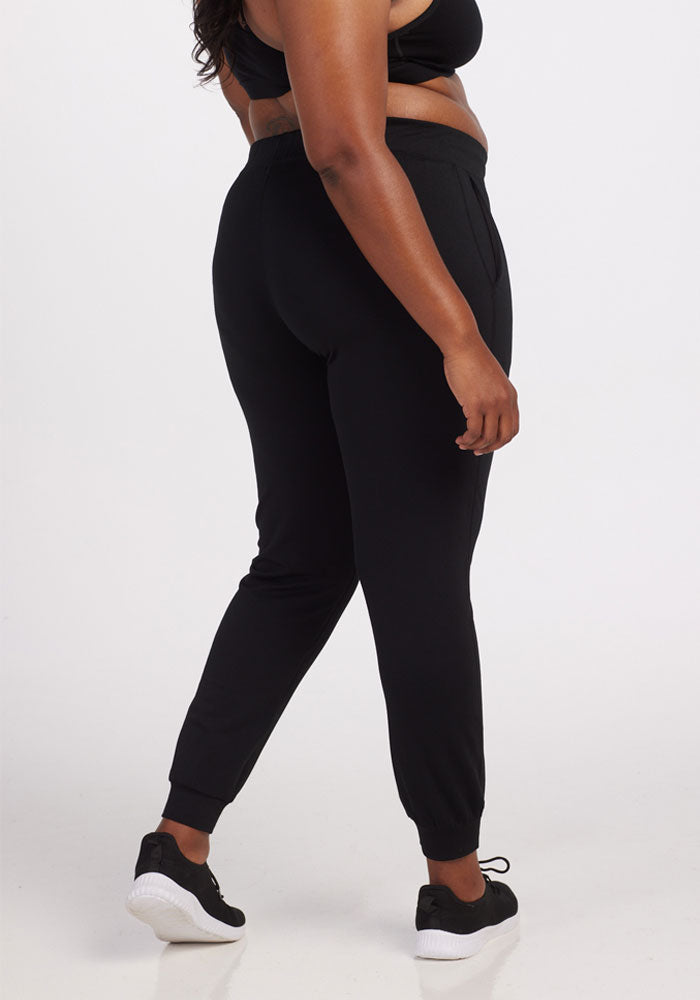 A person wearing a black sports bra, the Lola Jogger - Black from Woolx, and black sneakers with white soles stands against a plain white background. The person is facing slightly away from the camera, highlighting the travel-ready comfort and fit of the Woolx joggers.