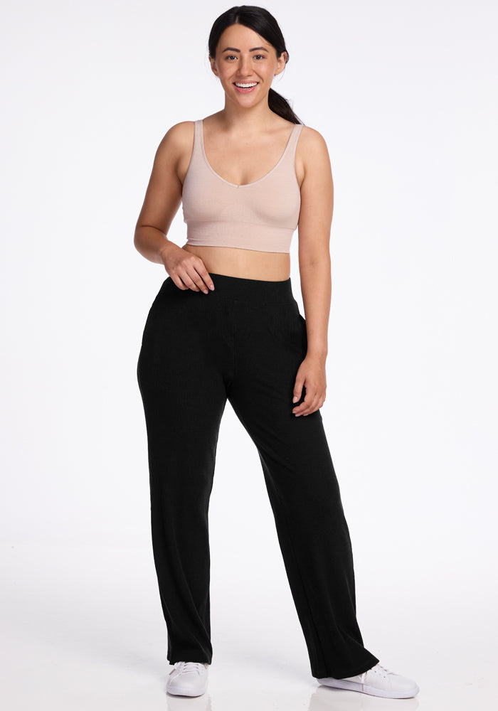 A person with long, dark hair is standing and smiling, dressed in a beige sleeveless crop top, Woolx's Luca Ribbed Pants in black, and white sneakers. The background is plain white, with the individual posing with one hand relaxed at their side and the other resting on their hip.