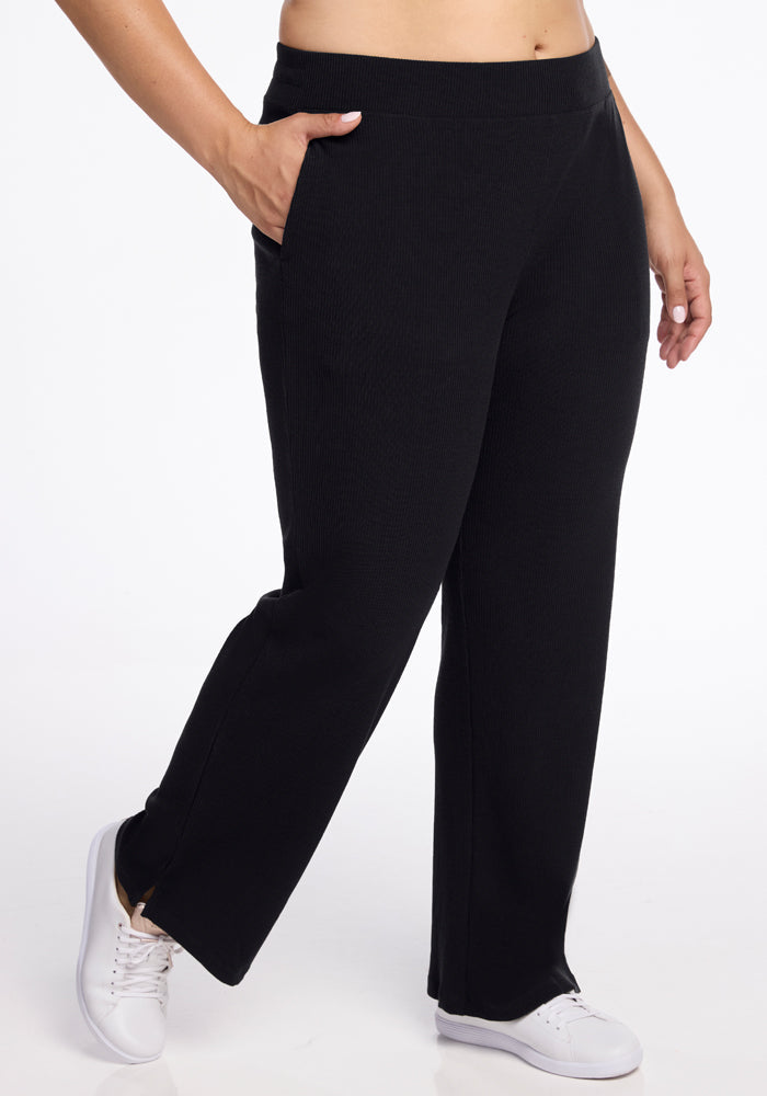 A person is seen from the waist down wearing black, straight-leg Luca Ribbed Pants from Woolx with hands in the pockets. The individual is also wearing white sneakers and is standing against a plain white background. | Juliana is 5'10", wearing a size XL