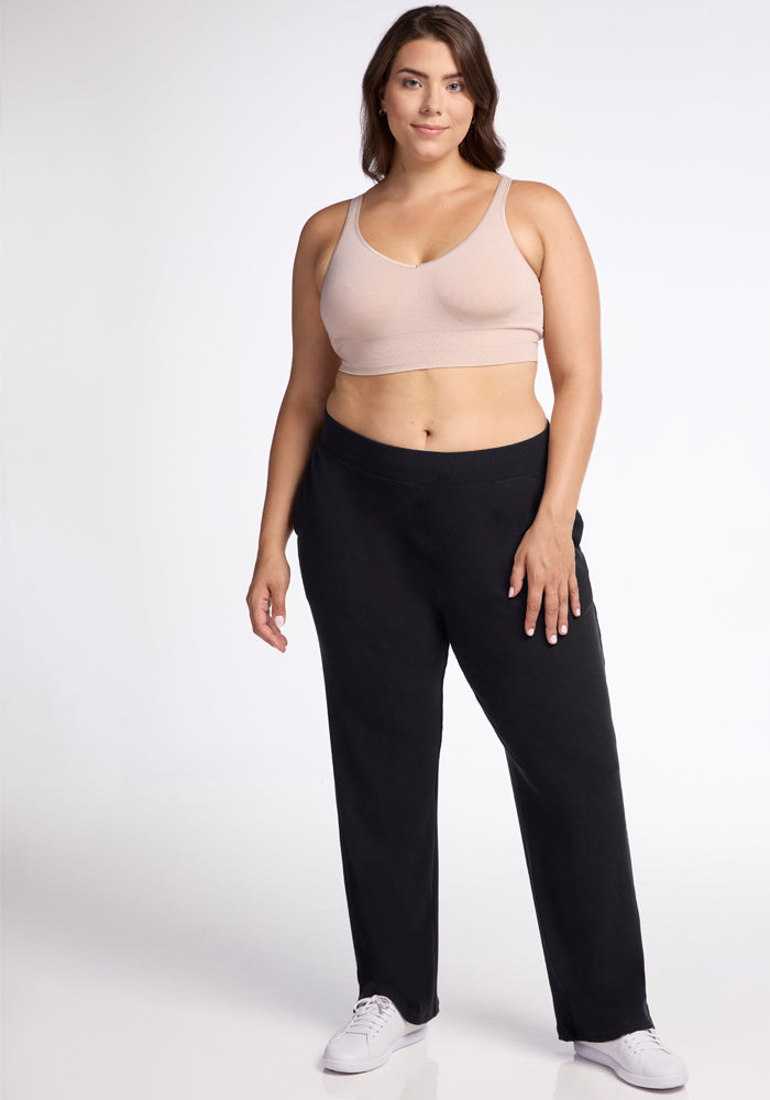 A woman stands against a white background, wearing a beige sports bra and the Woolx Luca Ribbed Pants in black with a wide-leg fit. She has shoulder-length brown hair and is smiling slightly. She is also wearing white sneakers.