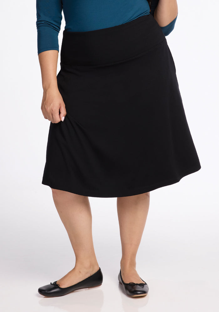 A person wearing a blue top and the Marilyn Flowy Midi Skirt in Black by Woolx, featuring its flowy design, stands on a white background. They are also sporting black ballet flats, with the image cropped from the shoulders down. | Juliana is 5'10", wearing a size XL
