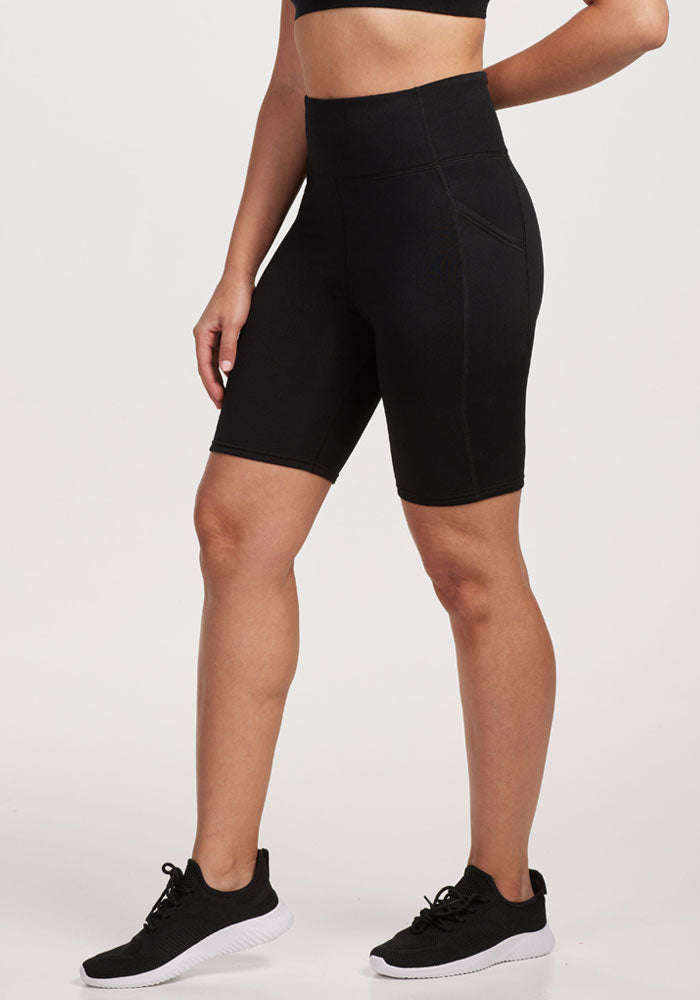 A person wears a black sports bra, Woolx Merino Flex® Dani Bike Shorts - Black with a supportive high waistband, and black athletic shoes, standing against a plain white background from mid-torso to feet.