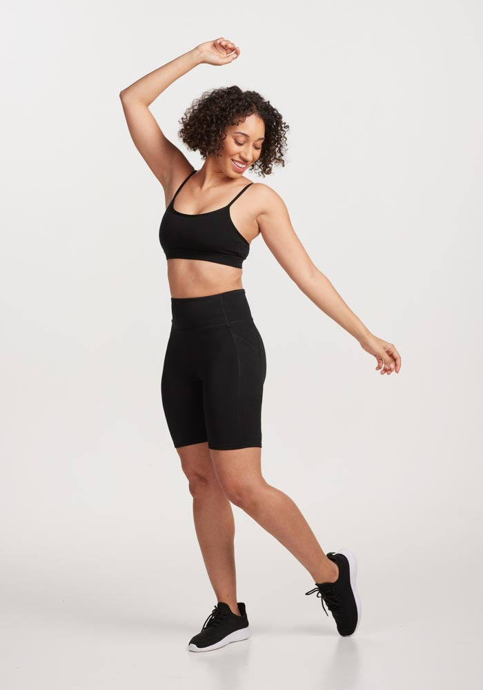 A woman in Woolx Merino Flex® Dani Bike Shorts - Black, a black sports bra, and black sneakers smiles with one arm raised against a plain white background.