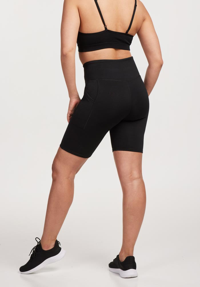 A person stands with one leg slightly bent, facing away from the camera against a white background, wearing a black sports bra, Woolx Merino Flex® Dani Bike Shorts in black with a supportive high waistband, and black athletic shoes.