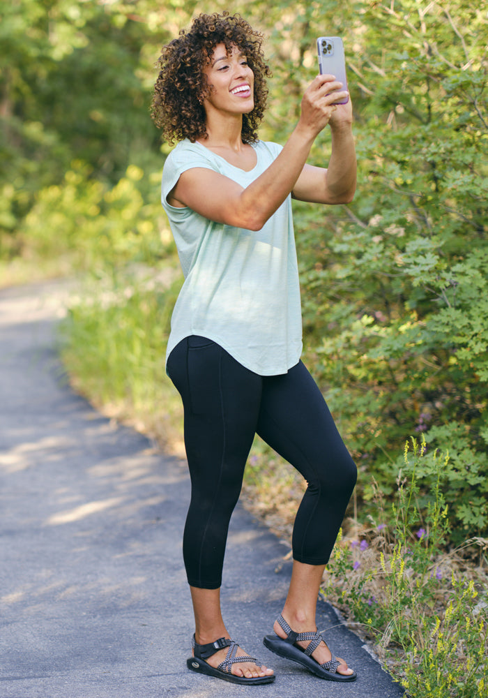 Smiling outdoors on a paved path, a woman snaps a selfie among lush greenery. She wears athleisure, including Woolx Merino Flex® McKenna Capris in Black, paired with a light green t-shirt and sandals.