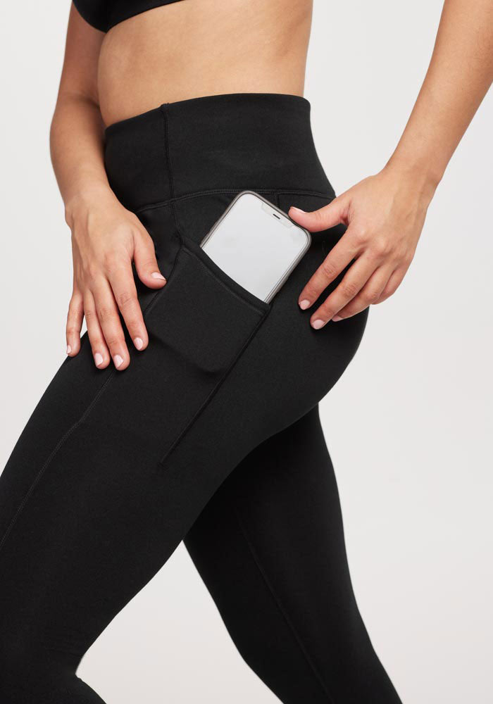 A person in Woolx Merino Flex® McKenna Capris - Black places a smartphone into the side pocket, with their midsection and upper legs visible against a light background.