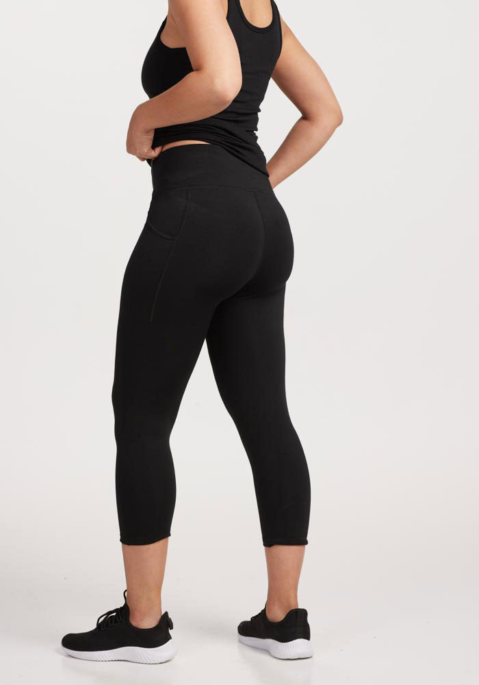 A person in black athleisure, featuring a tank top, Woolx Merino Flex® McKenna Capris - Black, and athletic shoes, stands facing away with hands on hips against a plain white background.