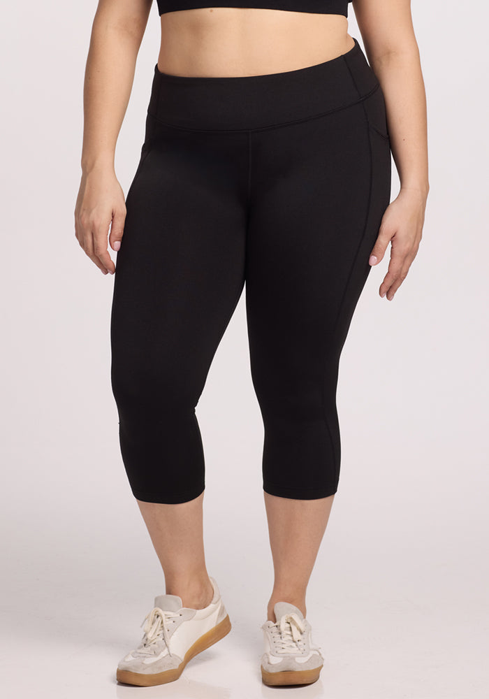 A person models the Woolx Merino Flex® McKenna Capris in black with a matching sports bra and white sneakers, standing against a plain light background with arms relaxed at their sides.