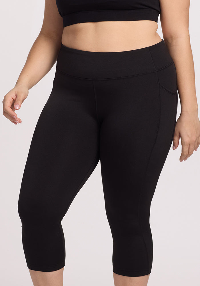 A person wearing Woolx Merino Flex® McKenna Capris in black and a crop top stands with one arm relaxed by their side against a plain light background.