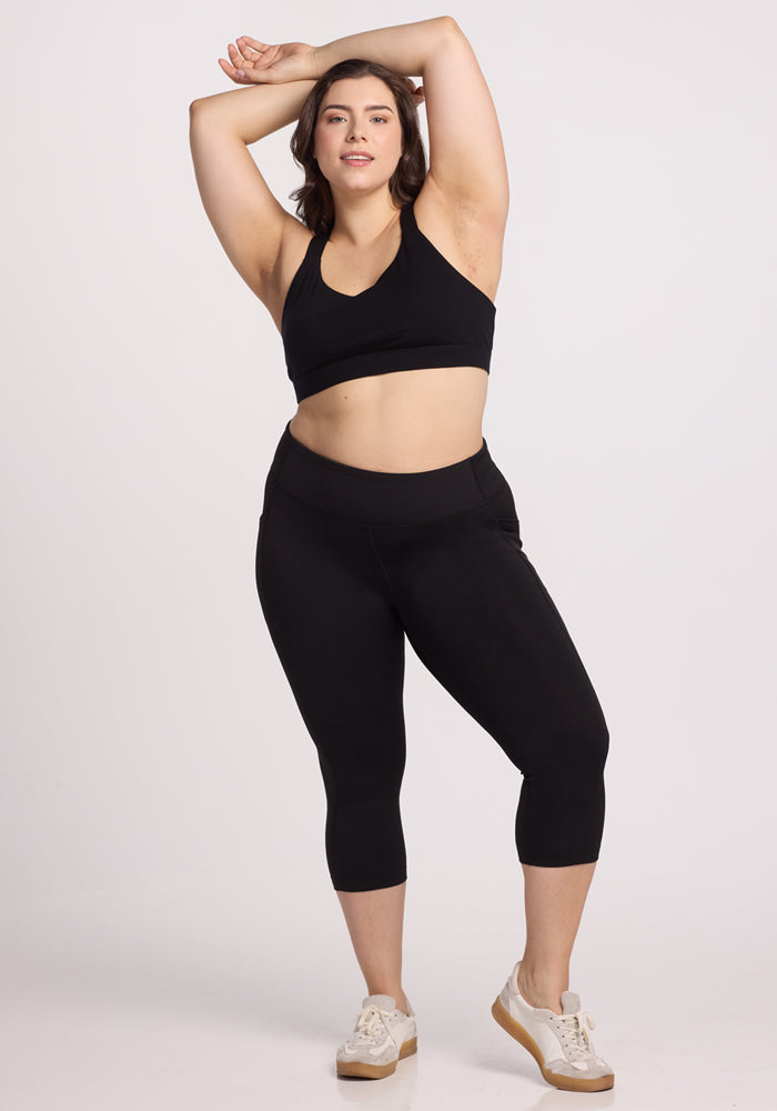 A woman models Woolx Merino Flex® McKenna Capris in black with white sneakers, posing confidently with one arm raised against a plain white background.