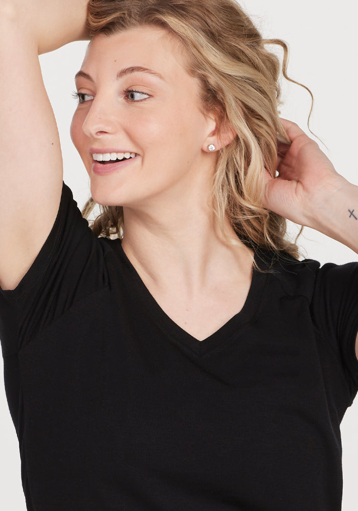 A person with wavy blonde hair smiles while looking to the side, wearing the high-quality Mia V Neck - Black shirt from Woolx. They have a small tattoo on their wrist and are holding their hair back with one hand. The background is plain and light-colored. , Black