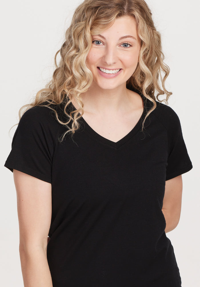 A person with long curly blonde hair is smiling, wearing the Woolx Mia V Neck in black, a high-quality women's T-shirt. They are standing against a plain white background., Black