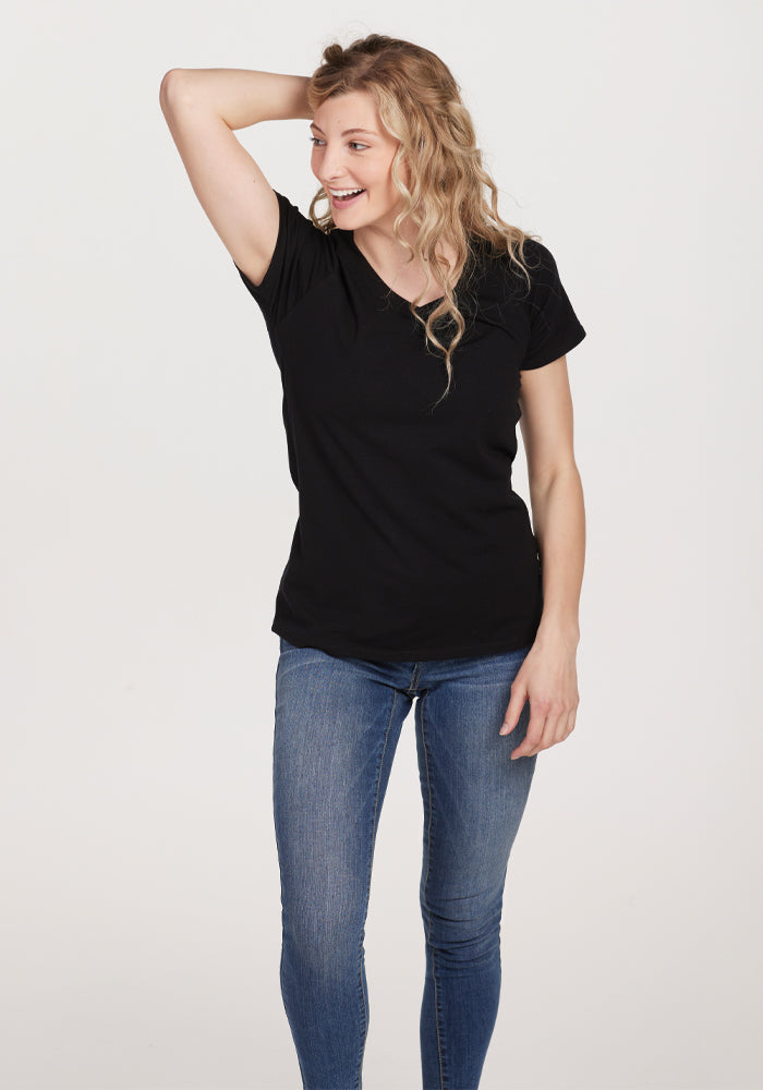 A person with long hair smiles, standing with one hand on their head, wearing the Mia V Neck in black by Woolx and blue jeans against a plain background. , Black