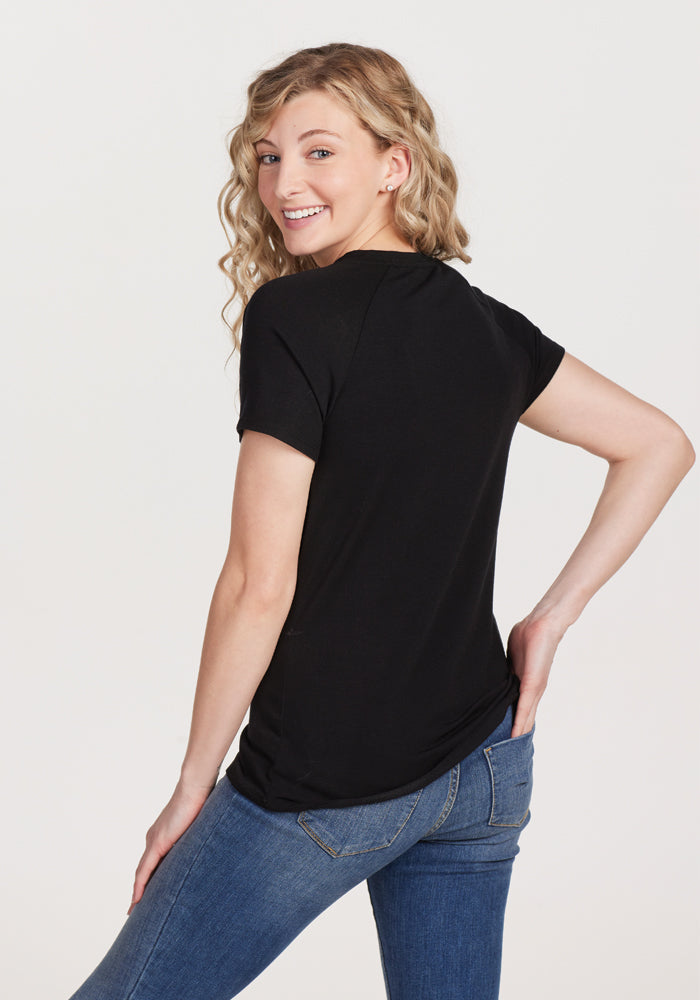 A person with long, wavy blonde hair is smiling and looking over their shoulder. They are wearing the Woolx Mia V Neck - Black, a superior quality women's merino wool tee, along with blue jeans, standing against a plain white background., Black