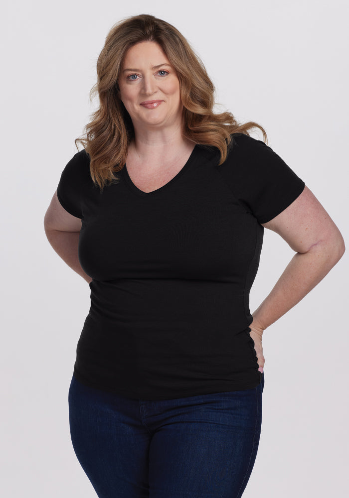 A person with long, wavy brown hair smiles slightly against a plain white background, wearing the Woolx Mia V Neck - Black in superior quality merino wool and dark jeans, hands on hips. | Cambre is 5'11", wearing a size XL