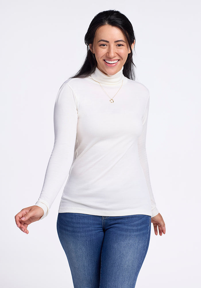 In a simple setting, a person smiles warmly in Woolx's Peyton Turtleneck in buttercream and blue jeans. Their long dark hair cascades over a delicate necklace, standing out against the plain white background. | Sarah is 5'8", wearing a size S