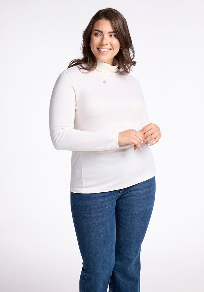 A person with long hair smiles against a plain background, wearing the Buttercream Peyton Turtleneck from Woolx made with breathable Merino wool, along with blue jeans. Their hands are gently clasped in front of them. | Juliana is 5'10", wearing a size XL