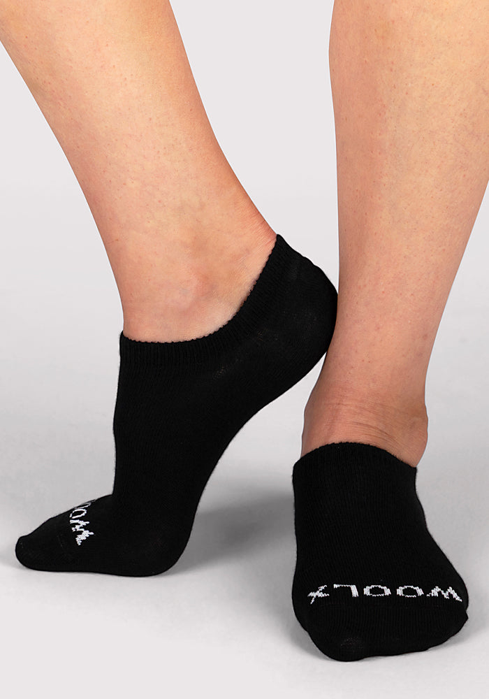Two feet wearing Woolx Womens Daisy Ped Socks in black, featuring white text on the toes, shown against a light gray background.