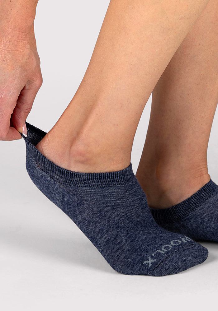 A person pulls up a Woolx Womens Daisy Ped Sock in Blue Jean Dream. The no-show women's ankle sock features a ribbed cuff and white text near the toes, shown against a plain, light-colored background. Final Sale item.