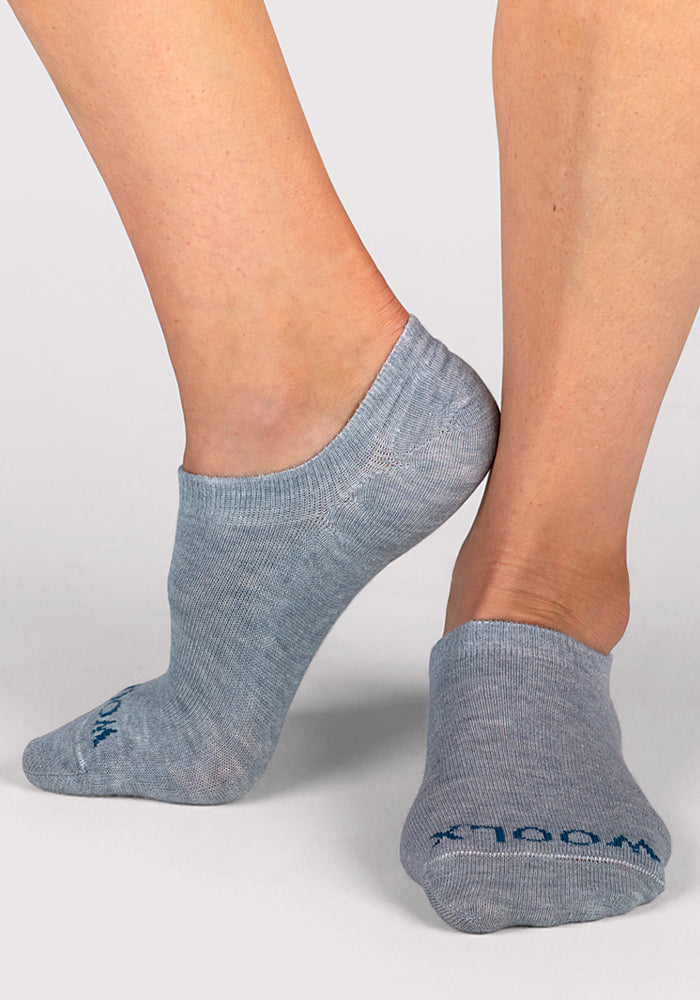 A person wearing Woolx Womens Daisy Ped Socks in Calm Waters (light gray Merino wool) stands on a light gray surface, with one foot flat and the other heel raised. Blue text appears on these women’s socks near the toes.