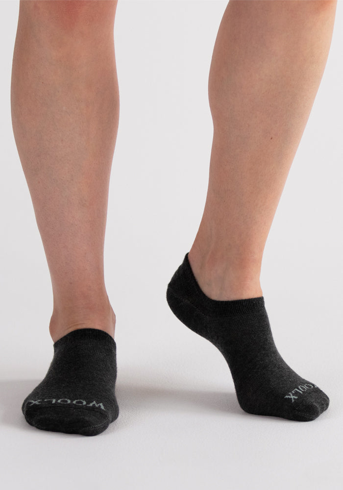 A person stands on a light surface wearing Woolx Daisy Ped Socks in Dark Grey Charcoal. One foot is flat, the other slightly raised, showing "WOOLX" in white letters on one sock.