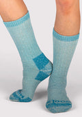 A person wearing Woolx Womens Extreme Crew Socks in Real Teal, featuring darker blue heels, toes, and toe lettering, stands on a light gray background with one foot flat and the other raised on the toes.
