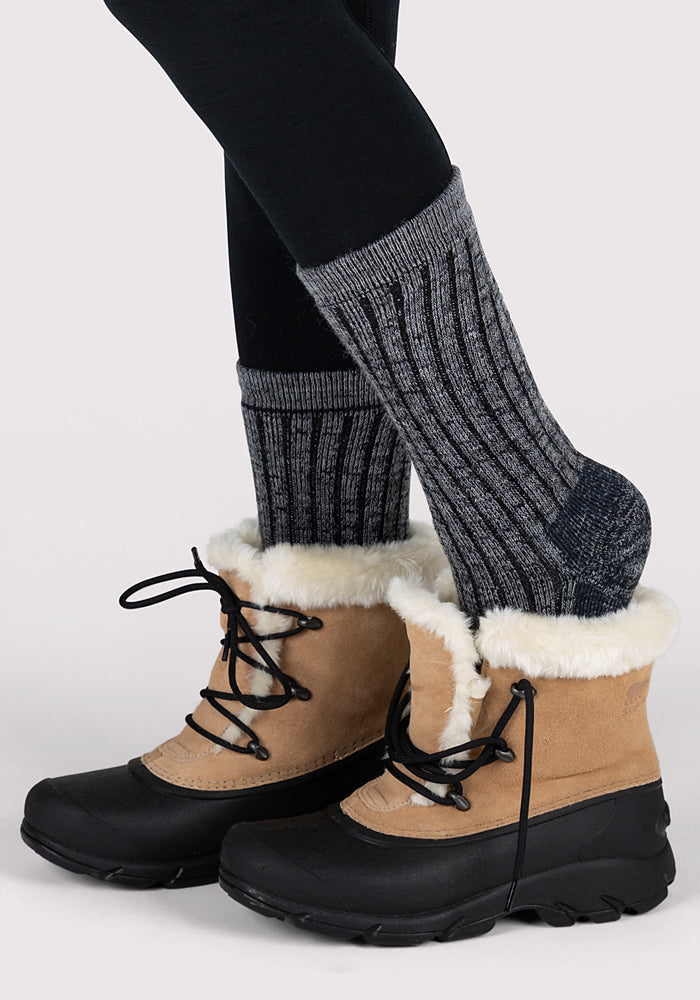A person wearing Woolx Womens Joey Crew Socks in Stone Grey and tan winter boots with black soles and white fur lining stands with one foot slightly ahead of the other against a plain background.