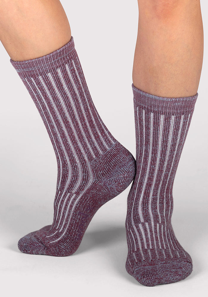 A woman wears Woolx Womens Joey Crew Socks - Wildflower, featuring purple and white ribbing, standing on a light grey background with one foot flat and the other slightly raised.