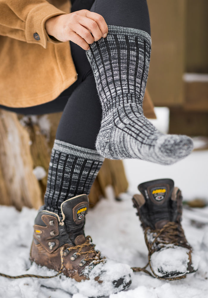A person perched on a tree stump slides on cozy and warm dark navy marled wool socks by Woolx over black leggings. The snowy terrain and hiking boots dusted with snow create an ideal winter outdoor setting for these snug Joey Socks, crafted from Merino wool.