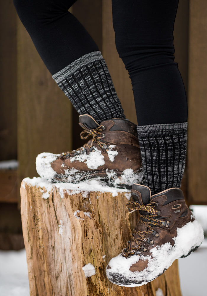 A person wearing cozy and warm brown hiking boots dusted with snow stands atop a snow-covered tree stump. Black leggings complement the Joey Socks - Dark Navy Marled by Woolx, adding warmth on this chilly day. Snow blankets the ground and wooden fence in the background.